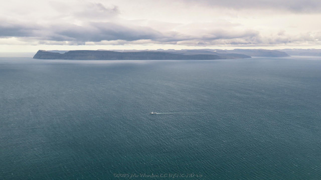 The view from the mountain top, across turquoise sea to a bare grey rock peninsula in the distance. A fishing boat is at sea midway between the viewpoint and the distant land. The sky is overcast and grey.