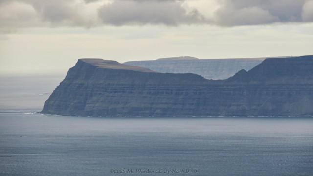 A close up photo of Grænahlíð, or the Green Cliffs, and specifically Rytur about 20 kilometres away which is a popular destination for hikers. Grey layered rocks are dropping almost vertically to the sea.