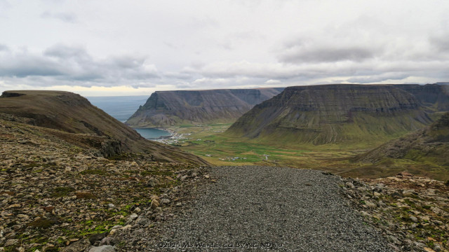 The view from a gravel path to the edge of the cliff top, looking inland from the mountain. A coastal town is visible far below. The pasture land surrounding the town is green and bordered by tall peaks with the turquoise sea in the distance.