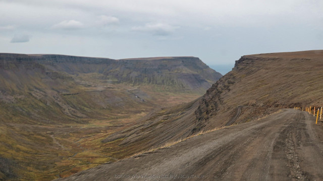 The drive down from Bolafjall to almost sea level. It's a dirt road with snow poles on the inside edge, and a sheer drop on the left down into a valley far below. A very tight hairpin bend is visible ahead, this has a short barrier on the outer edge. The valley beyond is U-shaped and glacial, clothed in autumnal colours. Waterfalls and a meandering river can be seen.