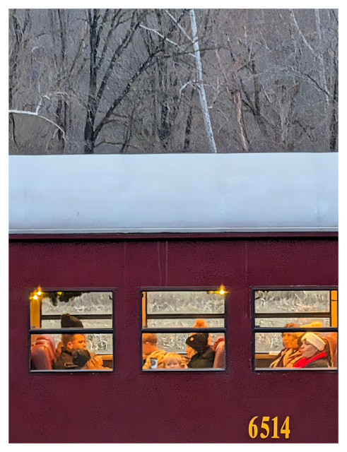 A maroon train car numbered 6514 is shown in profile. Through three lit windows, passengers in winter clothing sit closely together. A small child cups his hands to see out the window. Trees and bare branches fill the background, creating a cold, wintry scene outside the warm train interior.