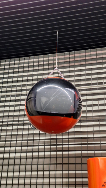 A spherical, red-orange TV set hanging on a chain in front of a metal lattice.