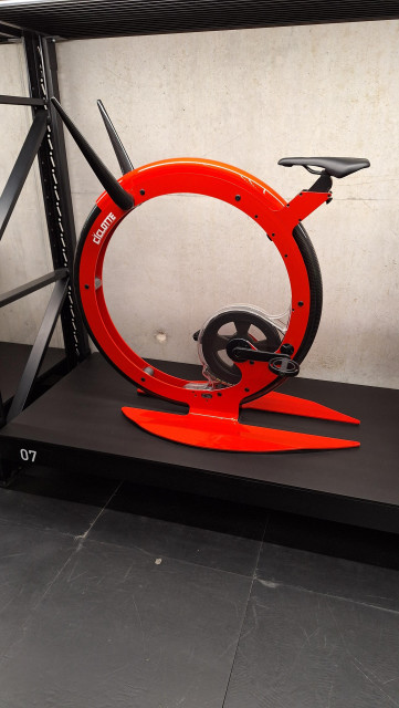 An orange-red, overdesigned treadmill bike. In essence one big wheel with duck feet, a saddle and handlebar sticking out the top. Pedals are stuck at the 4 o'clock position inside the wheel. It's displayed on a black shelf-like structure.