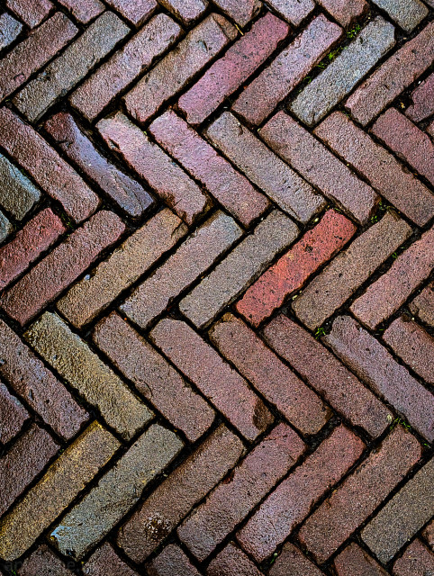 A brick paved surface laid in a herringbone pattern. They are made of clay, exhibiting a variety of natural, earthy tones, including reds, pinks, browns, and some muted grays or greens. 

The surface has a slightly rough, aged texture. There are visible gaps or joints between the bricks, filled with dirt, further defining the geometric pattern.