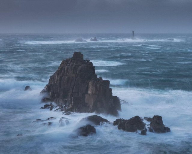 Ocean waves crash in a day gray cloudy sky against a shortish pillar of thick rocks.