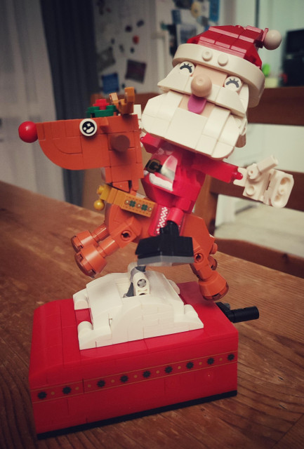 Figur aus Klemmbausteinen. Auf einem Rentier mit roter Nase sitzt ein fröhlicher Weihnachtsmann. Beide stehen auf einer roten Box aus Klemmbausteinen, in der eine Spieluhr mit Kurbel versteckt ist, sie spielt "Jingle Bells".
