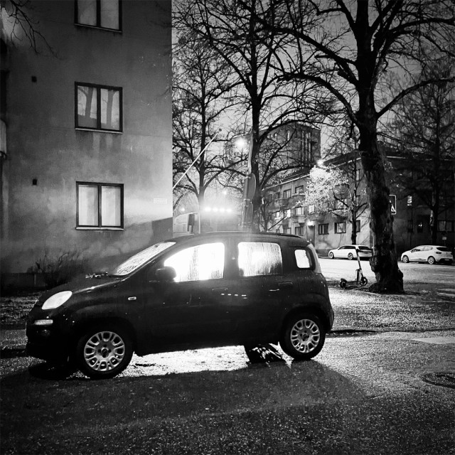 A black car parked on a dark wet street, backlit by a nearby street sweeper's lights, with dark trees and buildings in the background.