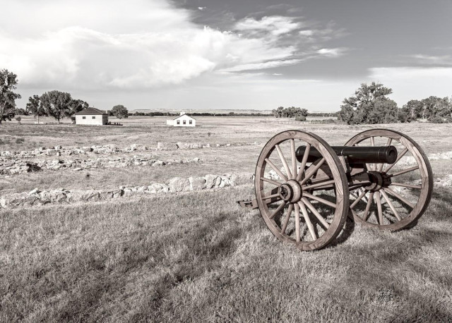 A black and white and slightly sepia toned image of a grassy field. There's a few trees on the far left and right in the distance. There are two small white buildings at center left. A restored 19th cannon on large wood wheels sits in the lower right pointing toward the right side of the frame.