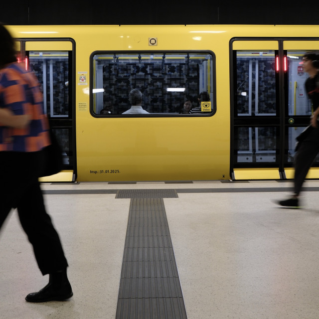 Das Foto zeigt eine kurzen Abschnitt eines gelben Berliner U-Bahnzugs, der an einem Bahnsteig hält. Links und rechts ist eine Tür, dazwischen ein Fenster. Die Türen der Bahn wurden gerade geschlossen - die roten LED-Leisten an den Türen leuchten. Durch das große Fenster in der Mitte sind im Inneren drei sitzende Personen zu erkennen, die mit dem Rücken zu den Fenstern sitzen. Der Boden des Bahnsteigs ist hell und mit dunklen tastbaren Leitstreifen versehen. Einer davon führt mittig vom Vordergrund zum Fenster des U-Bahnzugs. Der Blick wird dadurch auf die Person gelenkt, die fast mittig mit dem Rücken zum Fenster sitzt. 

Auf dem Bahnsteig bewegen sich zwei Personen von rechts nach links durchs Bild. Sie sind in Bewegung und dadurch sehr unscharf abgebildet. Die Person links ist im Vordergrund und schon teilweise außerhalb des Bildausschnitts. Die Person rechts ist im Hintergrund nah an der U-Bahn und betritt gerade den Bildausschnitt. Die Szene wirkt urban und alltäglich, aufgenommen in einer modernen U-Bahn-Station. Die dominante Farbe ist das kräftige Gelb des Zuges.


The photo shows a short section of a yellow Berlin subway train stopped at a platform. There is a door on the left and right, with a window in between. The train doors have just closed—the red LED strips on the doors are lit up. Through the large window in the middle, three people can be seen sitting inside with their backs to the windows. The platform floor is light-colored and has dark tactile guide strips. One of these leads from the center of the foreground to the subway train window. This draws the viewer's gaze to the person sitting almost in the center with their back to the window. 

Two people are moving across the platform from right to left. They are in motion and therefore appear very blurred. The person on the left is in the foreground and already partially outside the frame. The person on the right is in the background, close to the subway, and is just entering the frame. The scene appears urban and everyday, taken in a modern subway station. The dominant color is the bright yellow of the train.