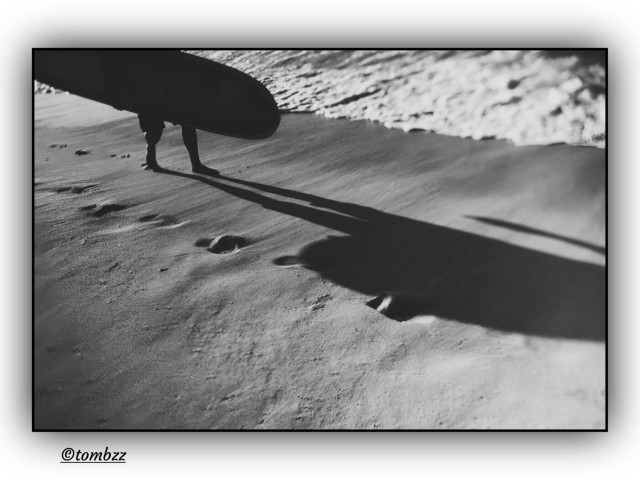 A black-and-white analog photograph shows part of a figure carrying a surfboard, with only the legs and the lower section of the board visible. The surfer walks along the sandy beach by the ocean, leaving clear footprints behind. The sun is at his back, making the shadow of the board and legs long and distinct, stretched across the grainy surface. In the background, foamy waves approach the shore. The framing is soft, with a slight vignette that gives the image a calm, contemplative mood.