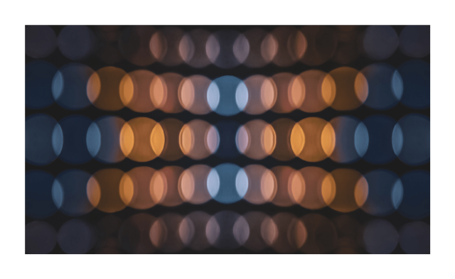 The photo is a pattern of blurred, out-of-focus circles arranged in rows and columns. The circles alternate in colour between warm, orange-toned hues and cooler, blue-toned hues. The background is dark, and emphasizes the glowing effect of the circles.