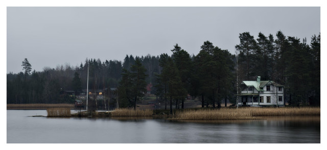 En liggande bild på ett vit trähus med ärgat, grönt tak på en udde med barrträd och vass. Bortanför lyser det i fönstren på ett annat hus.