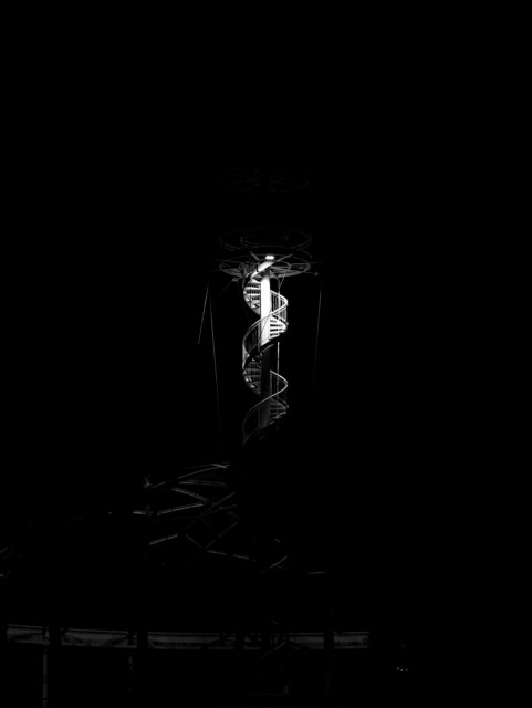 Black and white photo taken at night, so most of the image is black, with a bare white spiral staircase in the middle that fades into being around the brightly lit central column under what might be a circular platform. The stairs spin off leftwards into the dark below to meet unclear shapes glinting in the deep dark.