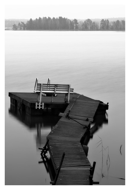 En stående svartvit bild på en brygga som sticker ut i vattnet. Längst ut står en vit bänk och två räcken för upp- och nedstigning i vattnet. I bakgrunden på andra sidan sjön är en skog.