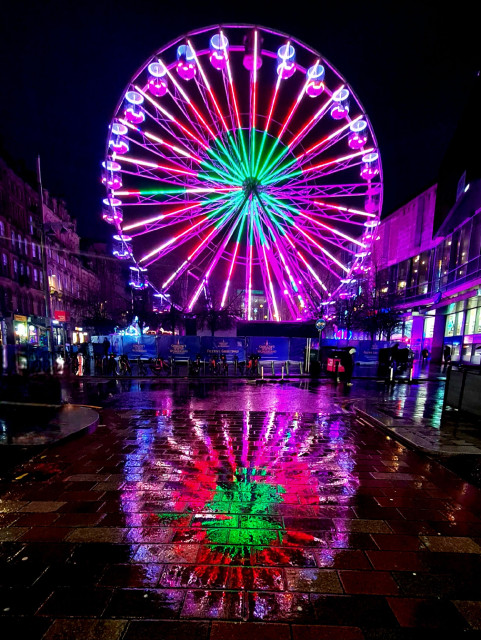 A ferris wheel at a Christmas market lit up at night and reflectes in the rain-soaked pavement.
