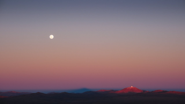 This photograph captures the mesmerising colours of an evening sky. The sky's gradient fades from blue at the top to white, then orange and pink at the bottom. A mountain range is visible at the bottom of the image, with all but one of the mountains already in darkness. The last illuminated one is on the right, with a metallic structure on top. In the middle of the mountain range, a triangular shadow can be seen. A silver full moon high in the sky on the left underlines the timelessness of the image.