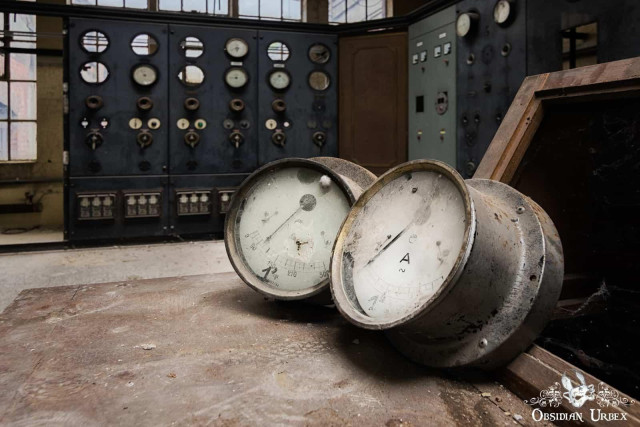 A photo of an abandoned industrial setting. Two large, dismounted gauges rest on a dirty surface in front of large control panels. 