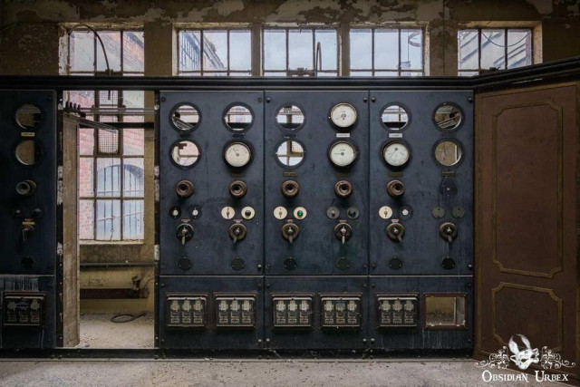 This image shows an old, dusty control panel with gauges, switches, and levers, suggesting an abandoned industrial setting. Sunlight streams through grimy windows, illuminating the decay. 
