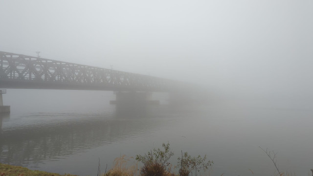 Eine Fachwerkbrücke führt über einen Fluß mitten in eine Nebelwand in der Bildmitte