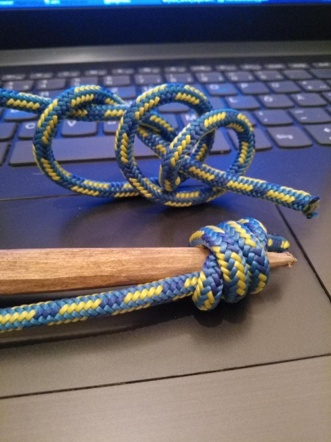 Ein zugespitztes Holz trägt einen Knoten, der sich zusammenzieht. Dahinter der lose gewundene Knoten in blau Reep, damit man die Konstruktion erkennen kann. Alles liegt auf einer grauen Laptoptastatur.