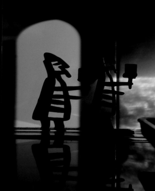 A picture of a small wrought iron candlestick resembling an old medieval guard in a helmet but only partially visible in the shadows on a wall. The candlestick's shadow is however fully visible and appears to be standing in a cellar vault under some old ancient castle. To the right of this are shadows from condensation running through a glass jug, making it look like reflections from the light from a stream are reflected on the wall above.