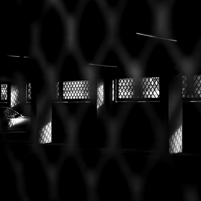 Black and white photo looking into a dark covered car park. Along the far wall, wide, glassless, windows are covered by diagonal grids. Light falls through these, cross-hatching sections of slim white pillars and the part of the front of a lone car facing the wall at the far left. The picture has been taken through similar bars, though these are blurred to the point of barely existing.