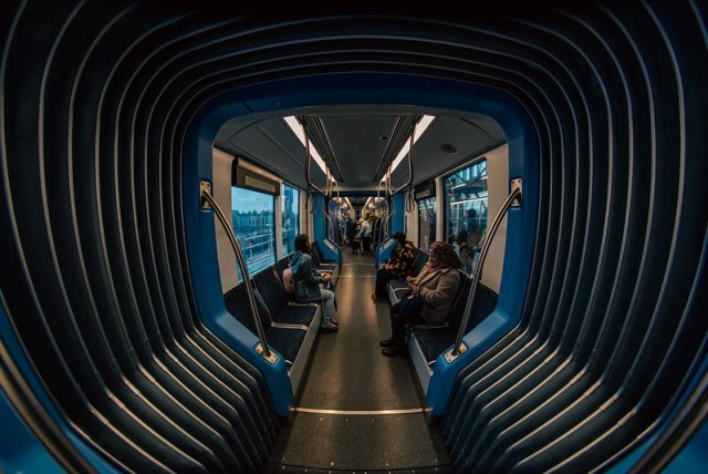 Fisheye shot looking down the length of a light rail train car. A short flexible section becomes a ribbed tunnel taking up half of the image, with the rest of the train car with people sitting and standing as they ride recedes into the far distance.