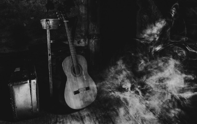 Man sieht einen Koffer einen Stuhl, an dem eine akustische Gitarre angelehnt steht. Von der rechten Seite strömt Rauch in das Bild hinein. Alles ist in Schwarzweiß und ziemlich dunkel gehalten.