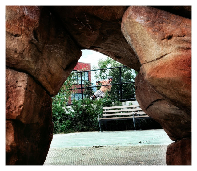 Photo taken at a skate park, from behind a rock sculpture. The rocks in the extreme foreground form an arched frame through which we see the rest of the scene in the distance: a park bench, some greenery and wire fencing, and behind that, a shirtless male skateboarder in a gray helmet, shorts, and kneepads in the middle of executing a move. Farther off are a tree and a red building under a clear sky.