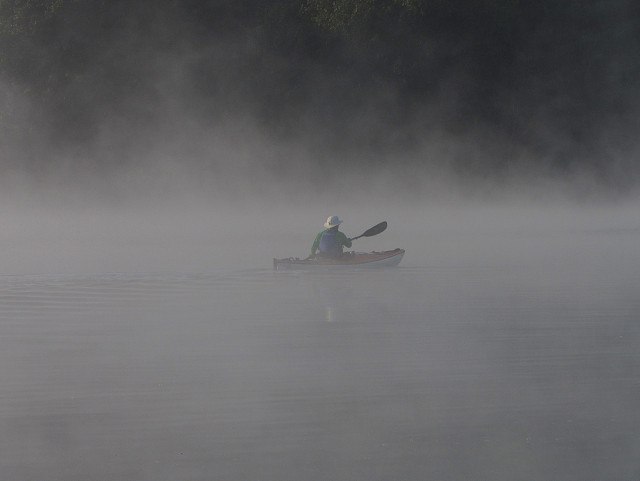 Eine Person mit weißer Mütze, grüner Jacke und blauer Schwimmweste paddelt in einem Kajak über einen nebelverhangenen See.

A person in white hat, green jacket, and blue life jacket, paddles a kayak across a misty lake. 