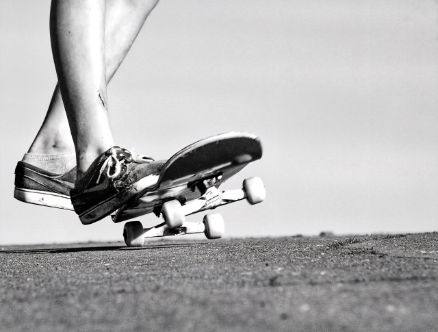 Das Bild zeigt in einer Schwarz-Weiß-Aufnahme die Beine und Füße einer Person, die auf einem Skateboard steht und sich offenbar fortbewegt. Der Fokus liegt auf der dynamischen Bewegung und den Details der abgenutzten Schuhe sowie des Skateboards. Die Szene spielt sich auf einem rauen, asphaltierten oder betonierten Untergrund ab, der im Vordergrund scharf abgebildet ist, während der Hintergrund unscharf und hell gehalten ist, was den Blick auf das Hauptmotiv lenkt. Die gekreuzten Beine und die Position des Boards vermitteln den Eindruck von Balance und Action.