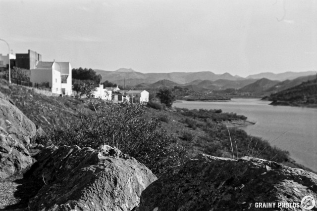 A black and white landscape featuring rocky terrain in the foreground, a calm lake with white houses on the left shore, and distant mountains under a clear sky. The scene captures a tranquil and natural atmosphere.