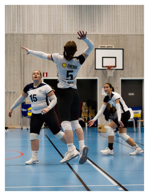 En stående bild från en match i volleyboll där en tjej hoppar upp med ryggen mot kameran för att smasha men bollen är inte i bild. Två lagkamrater fokuserar på bollen bredvid. De har marinblå byxor och vita tröjor.