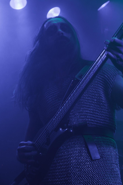 Ein Bassist mit Bart, langen Haaren und in Kettenhemd auf einer in Nebel gehüllter Bühne. Alles ist blau ausgeleuchtet