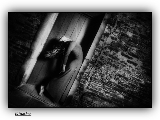 Black-and-white analog photograph in the style of expressive staged photography. A young woman in a long, simple dress with a fitted bodice and slightly flared skirt stands in a dramatic pose, leaning far back against wooden doors set within a stone frame. Her arms hang loosely at her sides, while her long hair falls straight downward, emphasizing the tension of the gesture. The background is a raw brick façade with a pronounced texture, marked by strong contrasts of light and shadow. The frame is tilted, intensifying the sense of movement and imbalance. The composition carries an artistic quality, with palpable tension between the solidity of the architecture and the expressiveness of the body. The atmosphere is dark, filled with ambiguity and emotional weight.