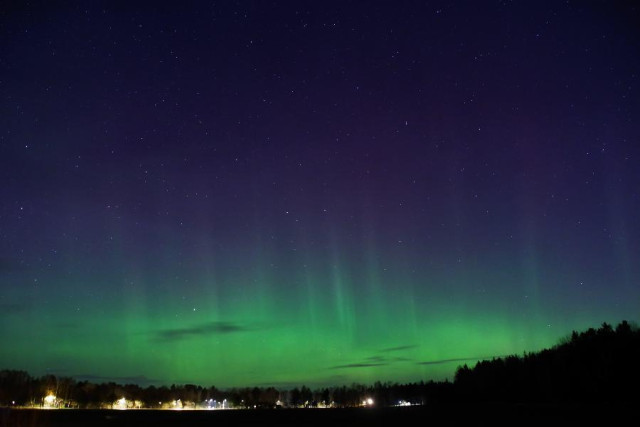 An image of aurora borealis on a starry sky above a field. Silhouettes of the forest to the right and trees following a street with street lights to the left. The aurora is pretty strong at the part nearest the horizon and above that some pillars reaching two thirds up and they are lilac at the top, blending to an almost white colour and below that one they are green. 