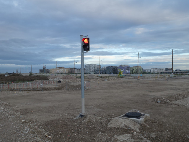 Mast mit Rotgelbampel mit leuchtendem Rotlicht steht in unbefestigter Baustellenbrache. Entfernt im Hintergrund Wohnhäuser eines Stadtentwicklungsgebiets