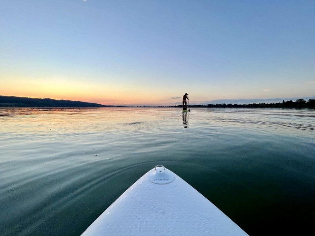 A paddleboarder stands on a calm body of water during sunset, with a soft gradient of colors in the sky and distant hills in the background. The foreground features the edge of a paddleboard.