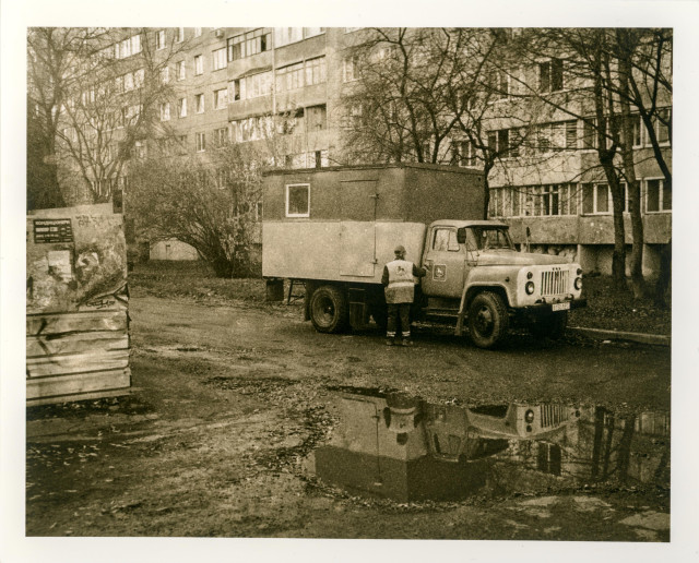 This lith print captures a quiet, slightly desolate residential courtyard, rendered in warm, gritty tones characteristic of the process. An old utility truck, its boxy form softened by the lith’s creamy highlights and coarse grain, stands parked near a puddle that mirrors the vehicle and the bare trees around it. A worker in a reflective vest leans against the truck’s side, adding a small but human focal point within the otherwise still, almost static scene.

The surrounding Soviet-era apartment blocks rise in the background, their weathered façades blending into the textured shadows produced by the lith development. The scattered leaves, the warped posters on a nearby metal structure, and the muted, fog-like contrast all contribute to a mood of quiet routine—an everyday moment transformed by the lith process into something nostalgic, atmospheric, and slightly melancholic