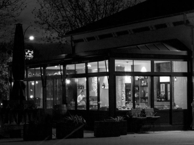 Une photo en noir et blanc, de nuit, format paysage, qui montre la structure légère d'un restaurant aux grandes baies vitrées éclairées de l'intérieur.  Une jeune femme lit ce qui ressemble à la carte du restaurant en attendant, peut-être, de prendre son service au milieu des tables dressées. Sur la placette devant le bâtiment  quelques tables et chaises de grandes jardinières fleuries, et sue la gauche, la silhouette d'un parasol replié.