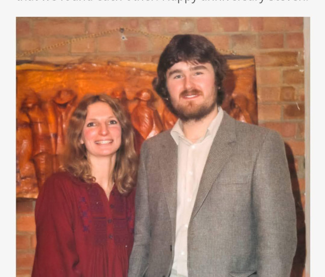 A smiling young couple standing in front of a word carving on a brick wall. The woman wearing an autumn red dress with blue embroidery has curling shoulder length hair. The young man has a beard, am earring, a lovely happy smile, lots of brown hair and a short beard with a moustache.