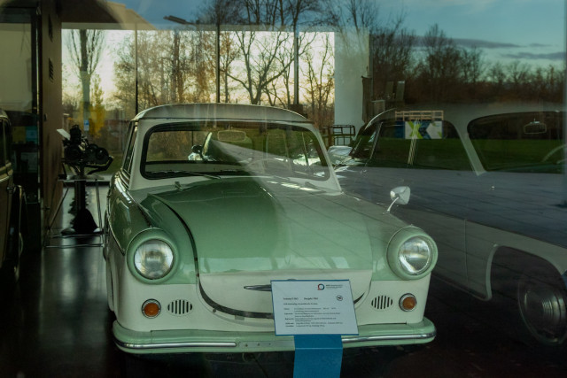 Ein Trabi 502 hinter einem großen Fenster mit Spiegelung des Parks hinter mir 