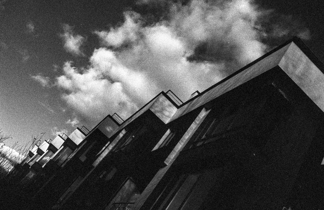 The image shows a row of modern, box-shaped buildings shot from a steep diagonal angle, giving the scene a dramatic, disorienting feel. The architecture forms a repeating zigzag pattern that leads the eye into the distance. Heavy film grain and deep contrast create a gritty, almost noir atmosphere. Above the buildings, large, textured clouds stretch across a dark sky, adding to the moody, cinematic tension of the composition.
