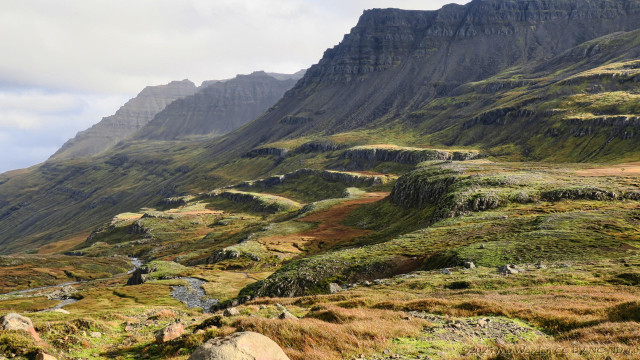 A colour photo of a mountainside sunlit from the right. The flank is divided into multiple small steps, each one top-lit by the strong sunlight. The stony terraces are covered in green and gold plants. The sky is overcast and the distant peaks of the range are misty.