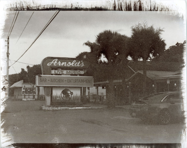 A neon sign has the word “Arnold’s” inside a curved arrow pointing toward a tropical styled building. Under the arrow it says “Live Music”, also in neon. Non-neon sign below that says “Bar • Kitchen • Entertainment”. There are palm trees in front of the building, and a car parked out front. Another sign behind is the kind with changeable letters and it says “Kitchen Open Daily. Dart Night. Fri Northern Exposure. Sat Sunjammer Band"