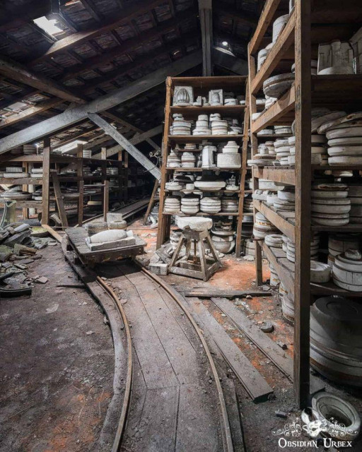 Dusty shelves filled with ceramic molds and pottery tools in an abandoned factory attic, with wooden tracks curving through debris on the floor.