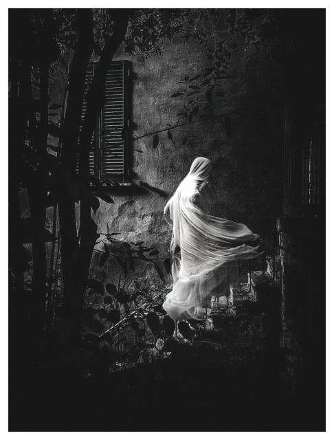 A black and white photo of a figure draped in flowing white cloth, set against the backdrop of a building outside. The figure is climbing up the staircase. The scene is partially obscured by trees and vegetation. There is a window with closed shutters is visible on the building in the background.