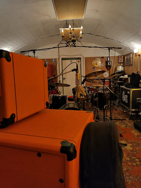 Photo of a mic'd drumkit, taken from behind a guitar amp stack. In front are two condenser mics in a Blumlein array.