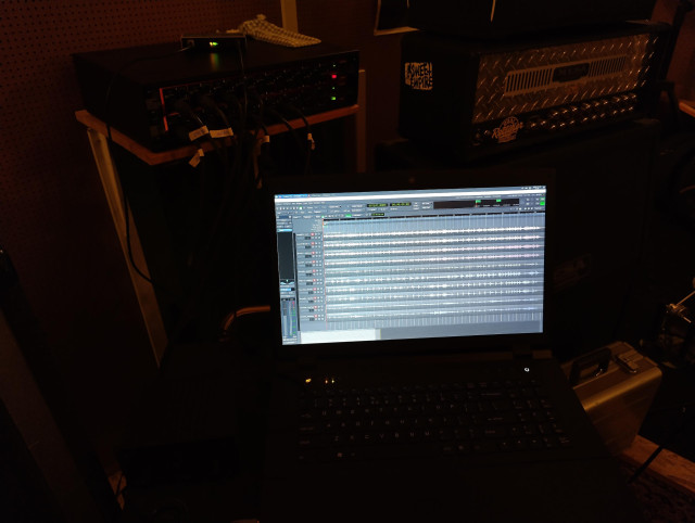 Photo of a bright laptop screen which shows Ardour with 9 simultaneously recorded tracks. In the back are the two ADAT units connected to the audio interface that provide 16 channels in.