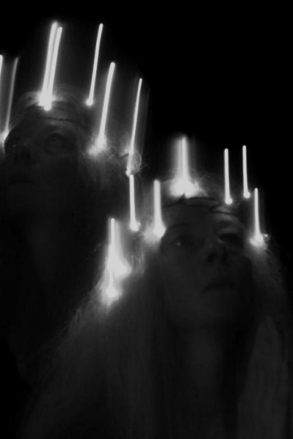 A vertical image in black and white with two faces of one woman's head and neck, seen obliquely from the front. The upper head is placed a little behind and above the right, and looks upwards and obliquely to the left. The lower head is placed lower right and slightly in front of the upper, and looks up obliquely to the right and holds up his palms in front of him. They both appear to have some kind of garland of candle-like lights around their heads, serious looks, and long hair. The image is an in-camera double exposure with ICM (intentional camera movement) in both exposures. 